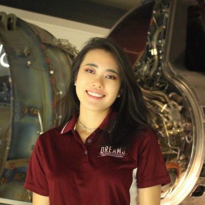 A person wearing a necklace and a maroon polo shirt with a logo saying DREAMS stands in front of a jet engine.