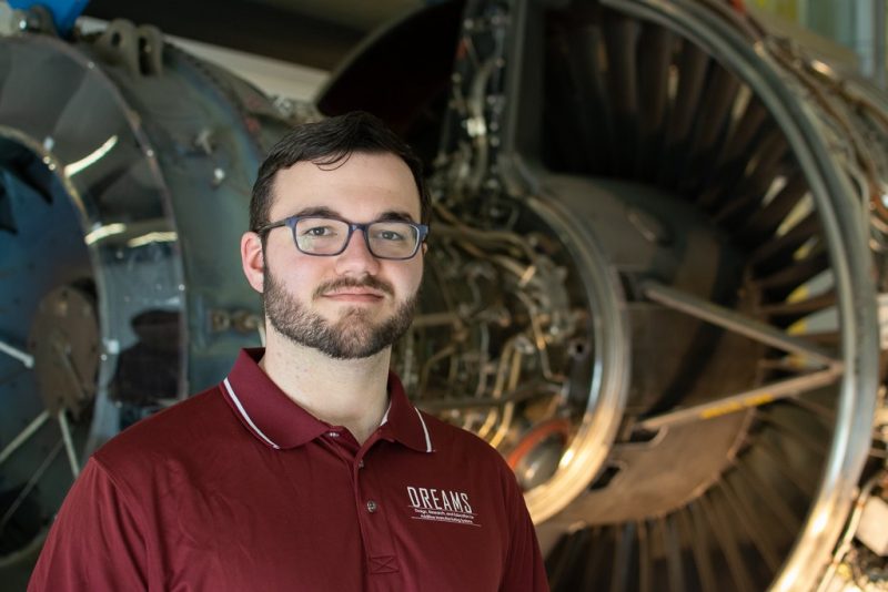 A person with glasses wearing a maroon polo shirt with a logo saying DREAMS stands in front of a jet engine.