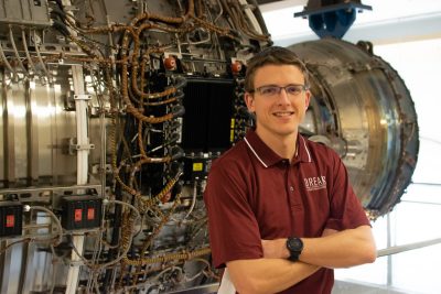 A person wearing a maroon polo shirt with a logo saying DREAMS and a black watch stands in front of a jet engine.