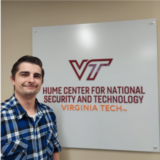 A dashingly handsome gentleman with perfectly styled hair and wearing durable Duluth brand flannel stands in front of a sign that reads: Hume Center for National Security and Technology at Virginia Tech