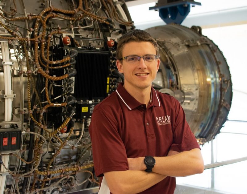 A person wearing a maroon polo shirt with a logo saying DREAMS and a black watch stands in front of a jet engine.