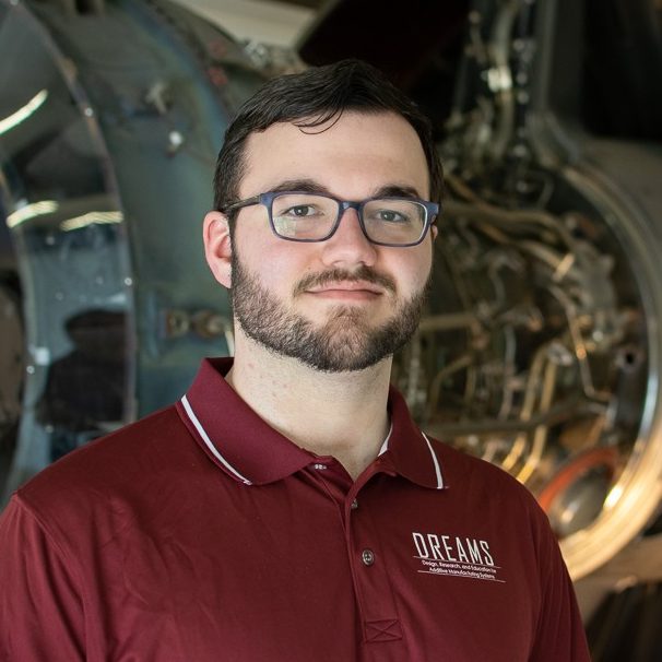 A person with glasses wearing a maroon polo shirt with a logo saying DREAMS stands in front of a jet engine.