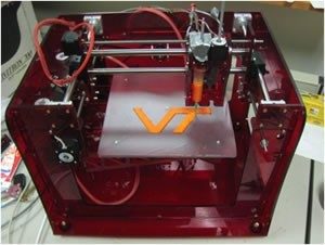 A 3-D printer in a cube shape. It is printing an orange V-T logo. The printer is mainly maroon.