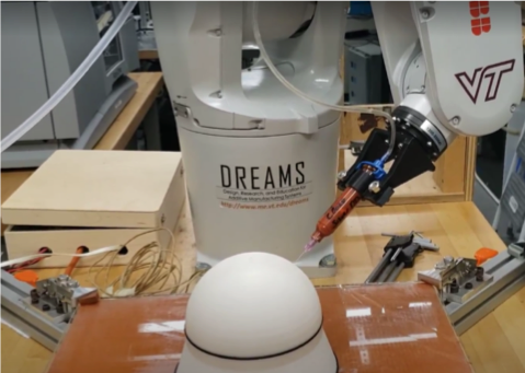 A small multi-axis robotic arm holds a syringe and prepares to conformally extrude material onto a dome